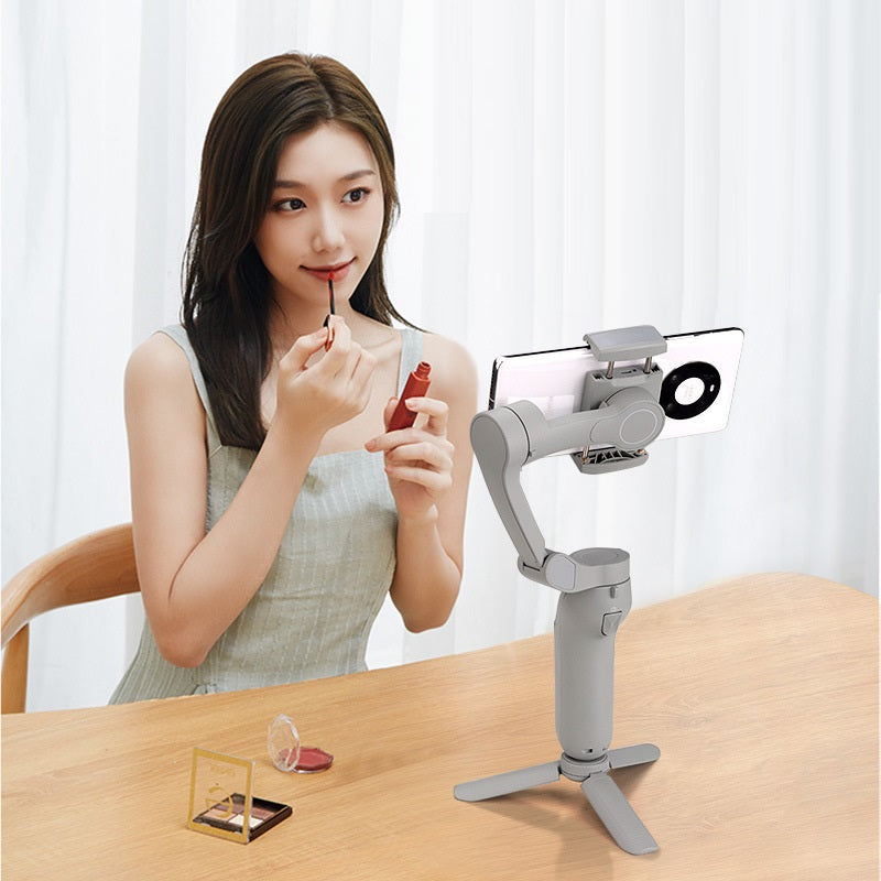Woman applying lipstick while recording with foldable 3-axis phone gimbal stabilizer on table