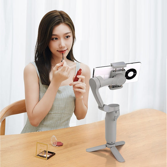 Woman applying lipstick while recording with foldable 3-axis phone gimbal stabilizer on table